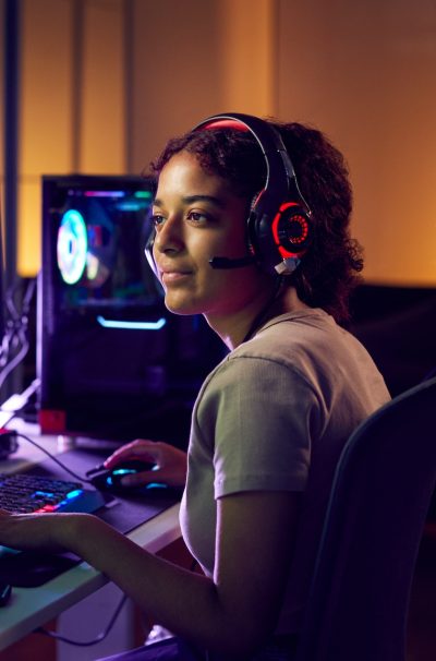 Teenage Girl Wearing Headset Gaming At Home Using Dual Computer Screens